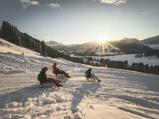 Hahnenköpfle: Ferienwohnung, Lodge und Hotel in Oberstdorf Drei Personen rodeln im Sonnenuntergang auf einem verschneiten Hang in den Bergen