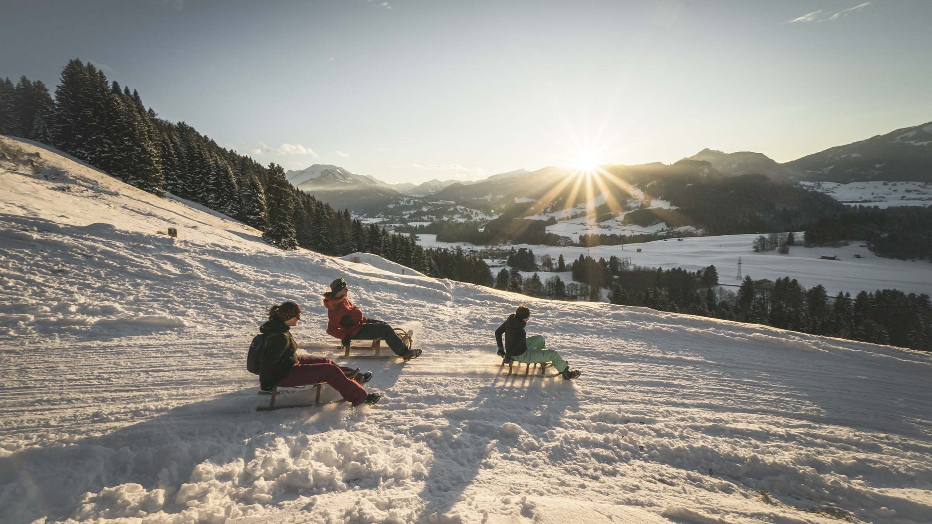 Plant euren Urlaub in Oberstdorf Drei Personen rodeln im Sonnenuntergang auf einem verschneiten Hang in den Bergen
