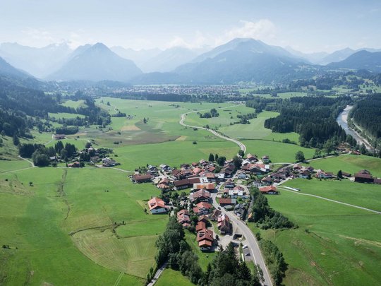 Plant euren Urlaub in Oberstdorf Luftaufnahme eines kleinen Dorfes umgeben von grünen Feldern und Bergen im Hintergrund