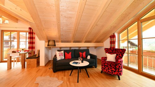 Your feel-good space at our hotel in Oberstdorf, Allgäu Cozy living room with wooden ceiling, sofa, red armchair, dining table, and large windows