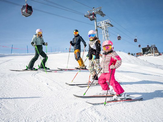 Hahnenköpfle: Ferienwohnung, Lodge und Hotel in Oberstdorf Familie mit Kindern beim Skifahren auf sonniger Piste mit Seilbahn im Hintergrund